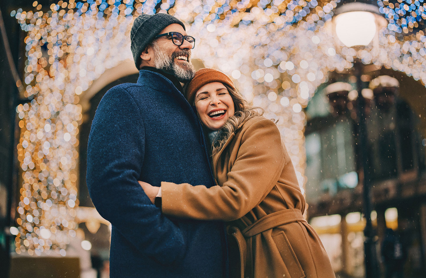 Ein älteres Paar steht vor einem glitzernden Hintergrund und umarmt sich. Beide tragen Winterkleidung und lächeln.