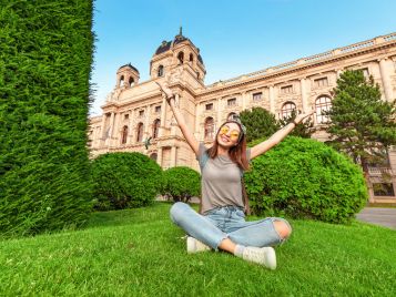 Eine junge Frau sitzt vor dem kunsthirstorischen Museum in Wien auf dem Gras im Schneidersitz und hebt erfreut die Hände in die Höhe.