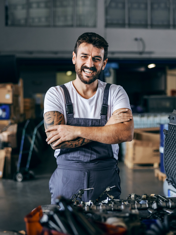 Ein Mann in Latzhose steht in einer Werkstatt und verschränkt lachend die Arme.
