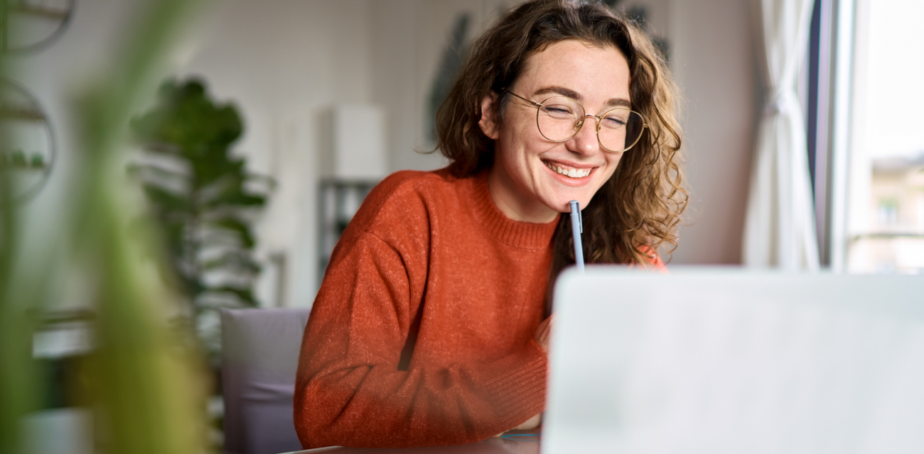 Eine junge Frau im orangenen Pullower sitzt lächelnd vor ihrem Laptop.