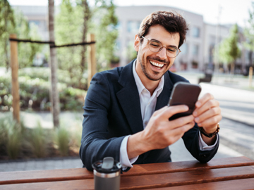 Ein Mann sitzt im Freien auf einer Bank und blickt mit einem breiten Lächeln auf sein Mobiltelefon.